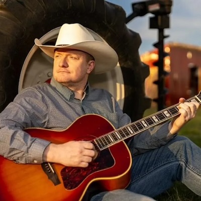 John Paycheck with guitar, sitting in front of tractor